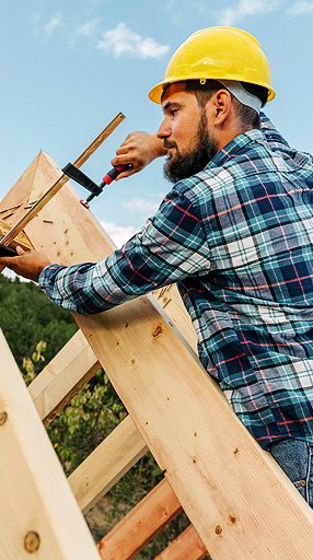 Construction worker in a yellow hard hat and plaid shirt using a clamp on wooden roofing materials, emphasizing precision and craftsmanship in roofing projects.