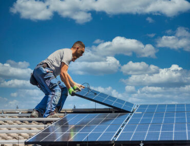Worker installing solar panels on a roof under a blue sky with clouds, emphasizing solar integration for Chandler homes by Ironclad Roofing.