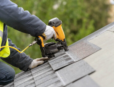 Roofing contractor installing asphalt shingles with a nail gun on a sloped roof, showcasing quality craftsmanship for Phoenix roofing services.