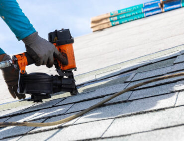 Worker using a nail gun to install asphalt shingles on a roof, with roofing materials visible in the background, representing Ironclad Roofing's expertise in roof replacement services in Phoenix.