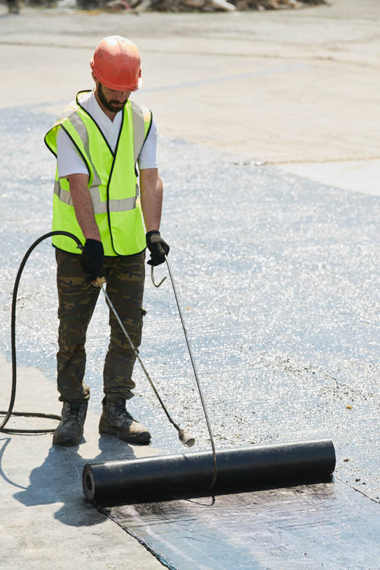 Worker applying rolled roofing material on a flat surface, wearing safety gear, including a hard hat and reflective vest, showcasing the installation process for durable roofing solutions.