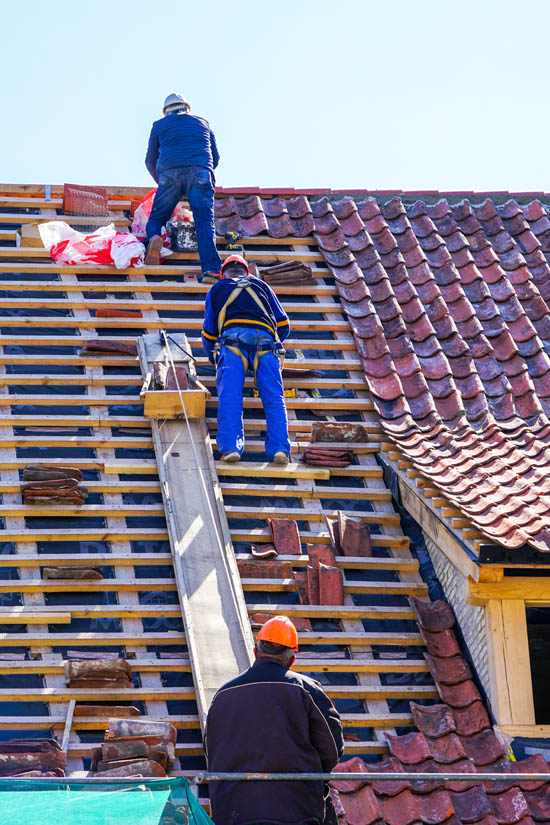 Roofing professionals installing tile on a sloped roof, showcasing a roof replacement process with safety equipment and scaffolding.