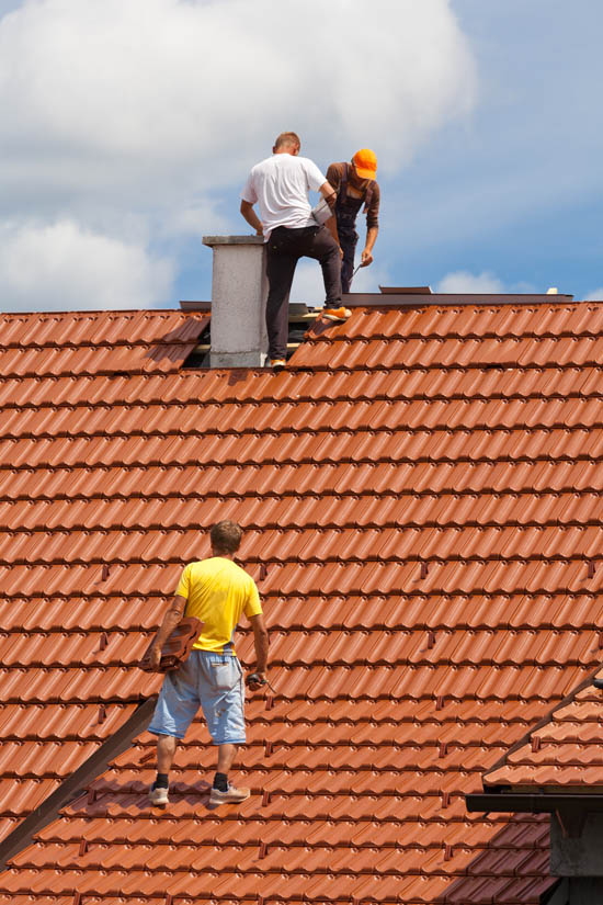 Workers installing tile roofing on a residential building, featuring a red clay tile roof under a blue sky with clouds, emphasizing professional roofing techniques.